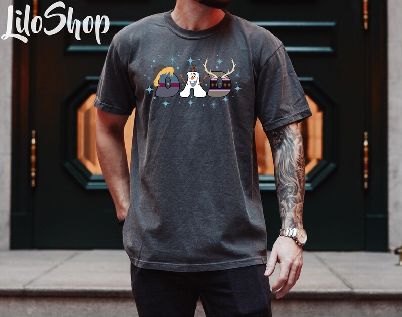 Puede incluir: Una camiseta gris oscuro con un gr&aacute;fico de los personajes de la pel&iacute;cula Frozen. El gr&aacute;fico muestra a Olaf, Elsa y Anna en un dise&ntilde;o estilizado. El texto "DAD" se deletrea usando las cabezas de los personajes.