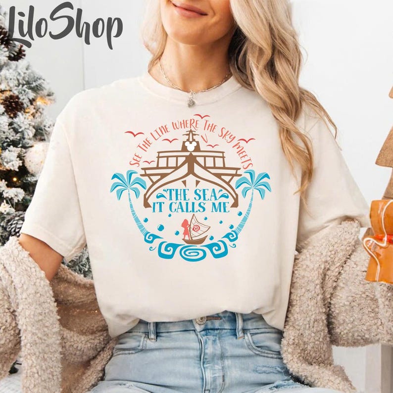 May include: Cream-colored t-shirt with a nautical design. The graphic features a ship, palm trees, and the text "SEE THE LINE WHERE THE SKY MEETS THE SEA IT CALLS ME" in blue and brown. The shirt is a relaxed fit.