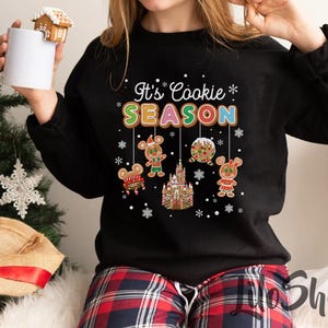 May include: Black sweatshirt with the text "It's Cookie Season" in colorful letters. The design includes gingerbread characters and a gingerbread castle. The person is holding a white mug with a gingerbread house on top.