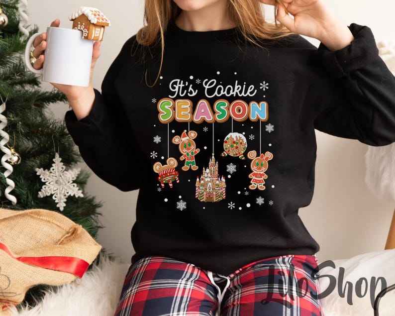 May include: Black sweatshirt with the text "It's Cookie Season" in colorful letters. The design includes gingerbread characters and a gingerbread castle. The person is holding a white mug with a gingerbread house on top.