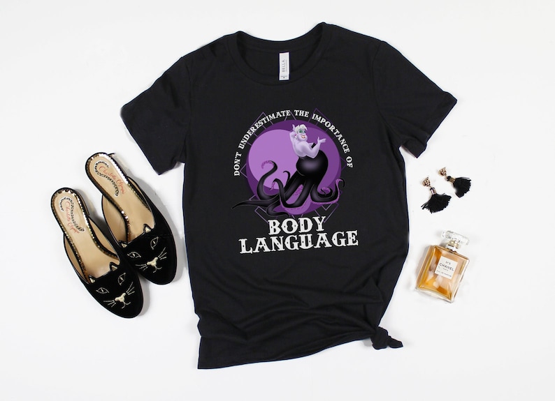 May include: Black t-shirt featuring a purple and black graphic of Ursula from The Little Mermaid. The graphic reads "Don't underestimate the importance of body language".