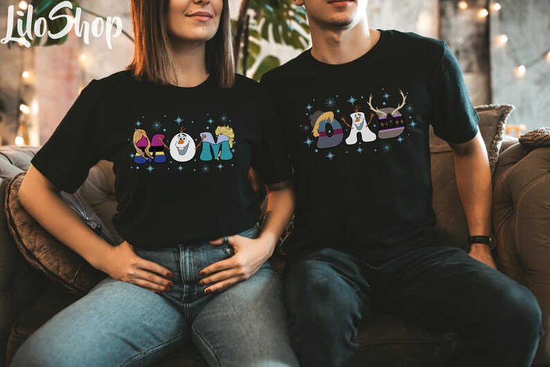 Puede incluir: Una pareja lleva camisetas negras con un dise&ntilde;o de Frozen. La camiseta de la mujer dice "MOM" con Olaf, Elsa y Anna. La camiseta del hombre dice "DAD" con Olaf, Sven y Kristoff.
