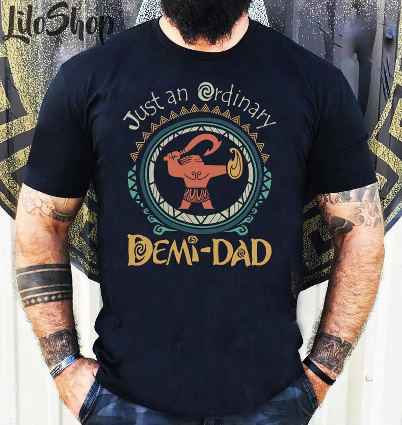 May include: Black t-shirt with the text "Just an Ordinary Demi-Dad" and a circular graphic featuring a cartoon figure holding a curved weapon. The graphic is in shades of orange, teal, and gold.