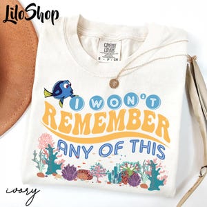 May include: Ivory-colored t-shirt with the phrase "I WON'T REMEMBER ANY OF THIS" in blue and orange, featuring a cartoon fish. The shirt has a crew neck and short sleeves. A gold necklace with a pendant is draped on the shirt.