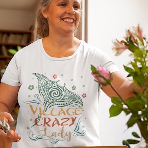 Puede incluir: Una camiseta blanca con un gr&aacute;fico verde y azul de una manta raya con el texto "Village Crazy Lady" en una fuente colorida.