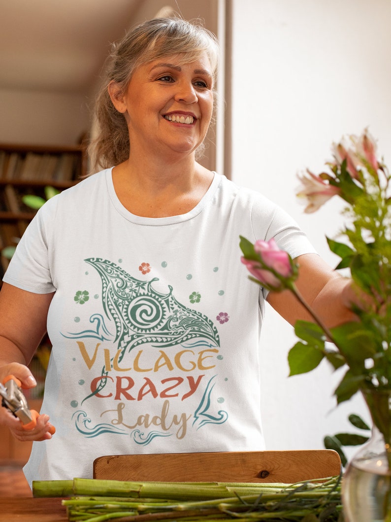 Puede incluir: Una camiseta blanca con un gr&aacute;fico verde y azul de una manta raya con el texto "Village Crazy Lady" en una fuente colorida.