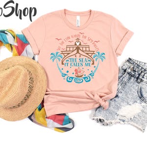 May include: A light pink t-shirt with the text "SEE THE LINE WHERE THE SKY MEETS THE SEA IT CALLS ME" and a graphic of a boat, palm trees, and waves. The shirt is paired with denim shorts, a straw hat, and a colorful scarf.