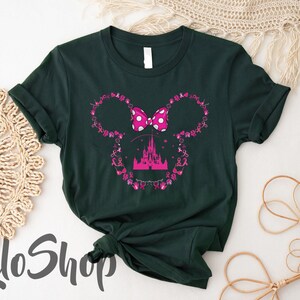 May include: A dark green short-sleeve t-shirt featuring a pink graphic of a castle silhouette centered within a mouse ear outline. The outline is composed of small pink breast cancer awareness ribbons and icons, topped with a pink polka-dot bow.