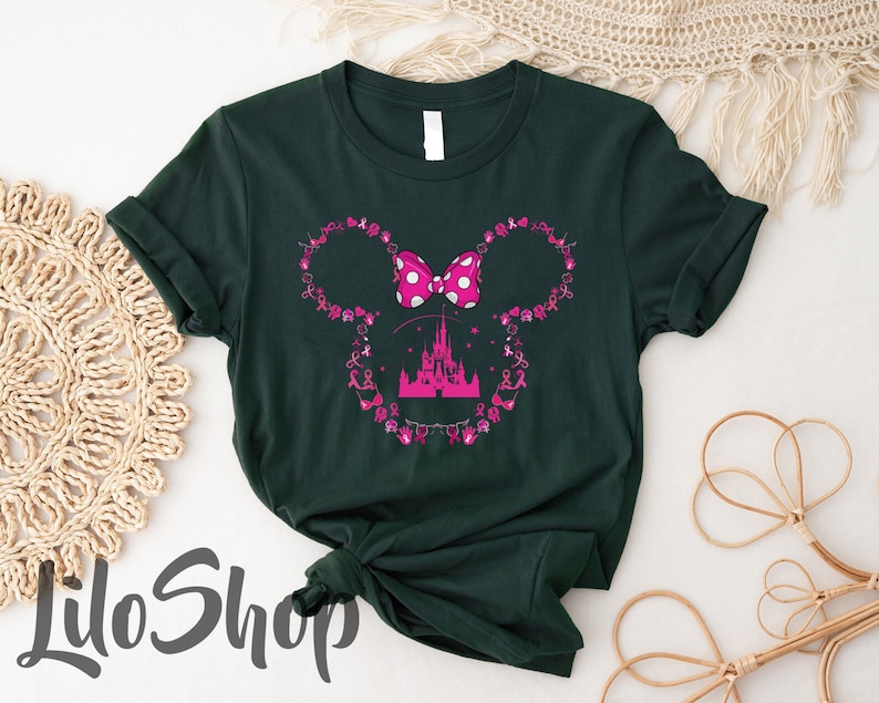 May include: A dark green short-sleeve t-shirt featuring a pink graphic of a castle silhouette centered within a mouse ear outline. The outline is composed of small pink breast cancer awareness ribbons and icons, topped with a pink polka-dot bow.