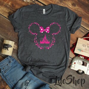 May include: A dark heather gray short-sleeve t-shirt featuring a graphic of a mouse-ear silhouette outlined with small pink breast cancer awareness ribbons and hearts. A pink polka-dot bow sits at the top, and a pink castle is in the center.