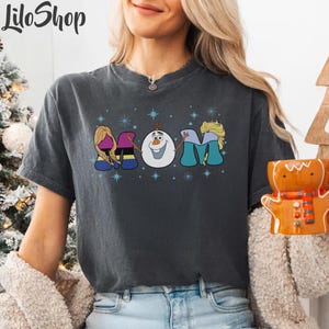 Puede incluir: Una camiseta gris oscuro con un gr&aacute;fico de los personajes de Frozen deletreando "MOM" con copos de nieve en el fondo.
