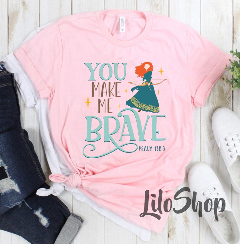 Może przedstawiać: R&oacute;żowa koszulka z grafiką przedstawiającą rudowłosą kobietę z łukiem i strzałami. Tekst na koszulce brzmi "You Make Me Brave" z odniesieniem do Pisma Świętego "Psalm 138:3".