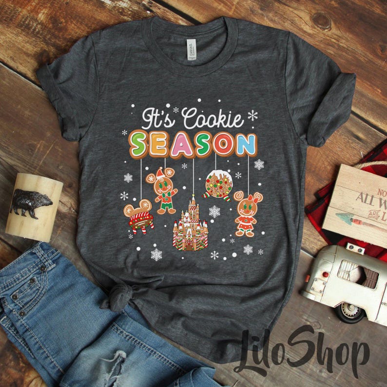 May include: Dark gray t-shirt with the text "It's Cookie Season" in colorful letters. The shirt features gingerbread cookie characters and a gingerbread castle design, perfect for the holiday season.
