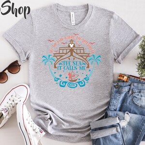 May include: A heather gray t-shirt with a nautical design featuring a ship, palm trees, and the text "The Sea It Calls Me." The shirt is paired with blue jeans, white sneakers, and sunglasses.
