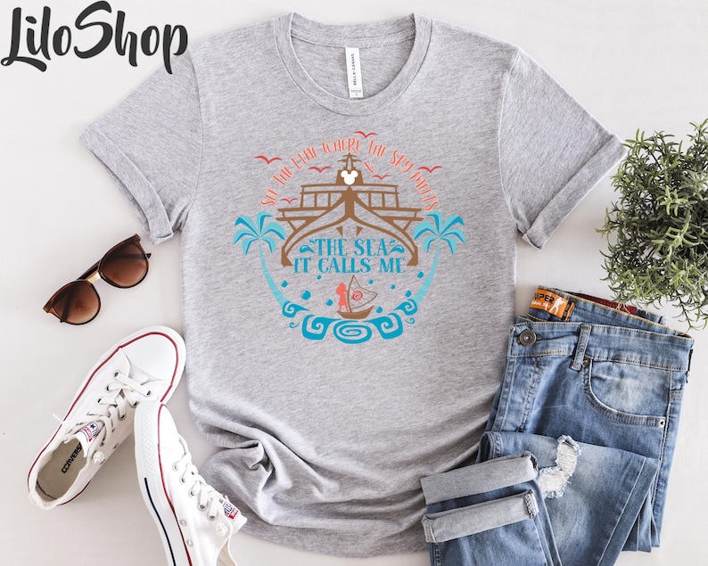 May include: A heather gray t-shirt with a nautical design featuring a ship, palm trees, and the text "The Sea It Calls Me." The shirt is paired with blue jeans, white sneakers, and sunglasses.