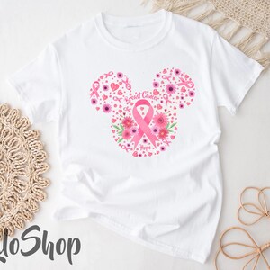 May include: A white short-sleeve t-shirt featuring a graphic design of a mouse-ear silhouette composed of pink flowers, small hearts, and breast cancer awareness ribbons, with the text Breast Cancer Hope printed in the center of the design.