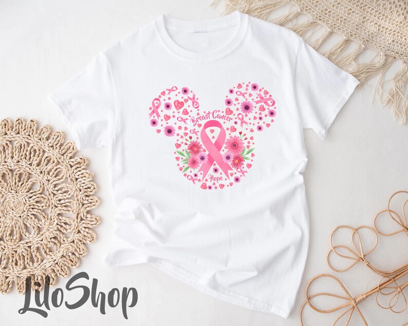 May include: A white short-sleeve t-shirt featuring a graphic design of a mouse-ear silhouette composed of pink flowers, small hearts, and breast cancer awareness ribbons, with the text Breast Cancer Hope printed in the center of the design.