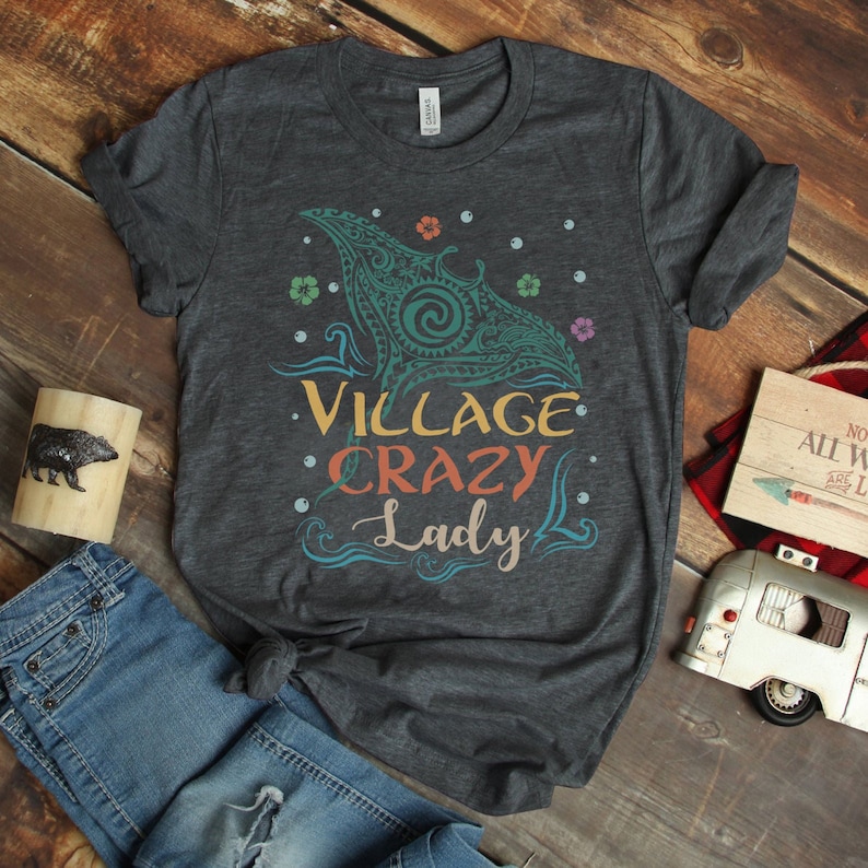 Puede incluir: Una camiseta gris oscura con un gr&aacute;fico colorido de una raya manta estilizada con el texto "Village Crazy Lady" impreso en ella.