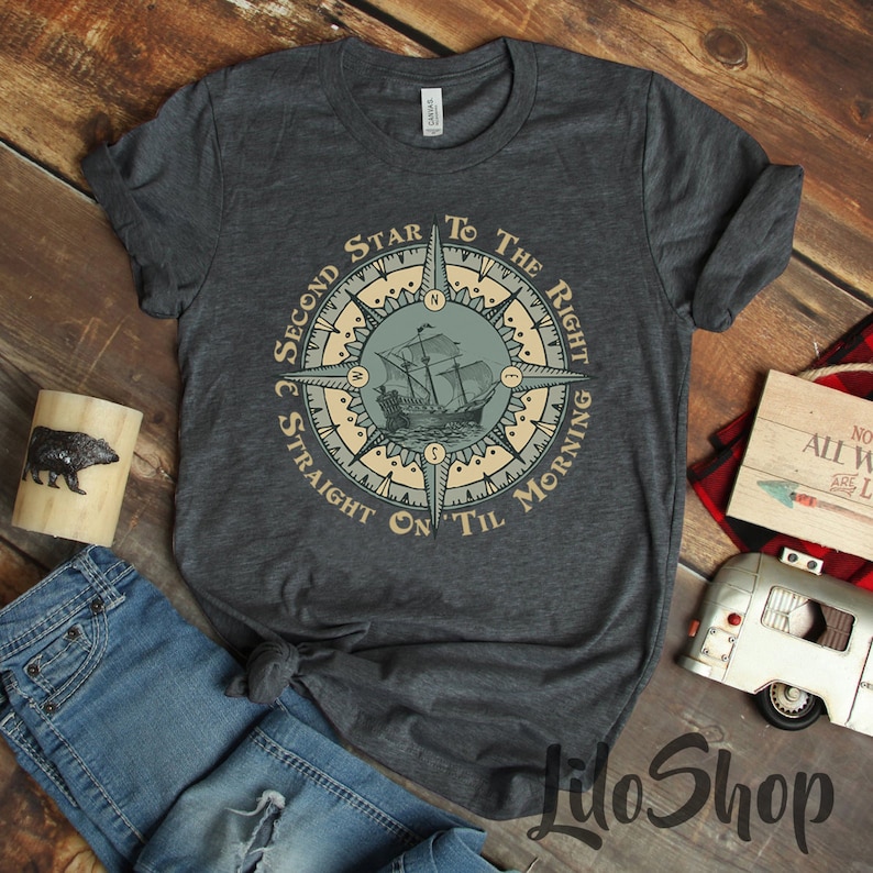 Puede incluir: Una camiseta gris oscuro con un dise&ntilde;o de br&uacute;jula que presenta un barco y el texto "Second Star to the Right Straight On Til Morning".