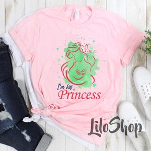 Puede incluir: Camiseta rosa claro con un dise&ntilde;o de sirena verde y roja y el texto "I'm his. Princess". La camiseta tiene cuello redondo y mangas cortas. El nombre de la marca "LiloShop" est&aacute; impreso en la parte inferior.