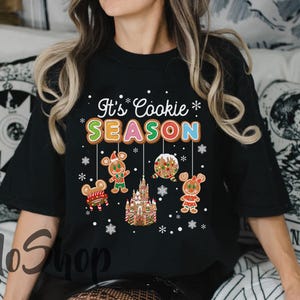May include: Black t-shirt with the text "It's Cookie Season" in white and colorful letters. The shirt features gingerbread cookie designs, including a gingerbread house, gingerbread people, and snowflakes.