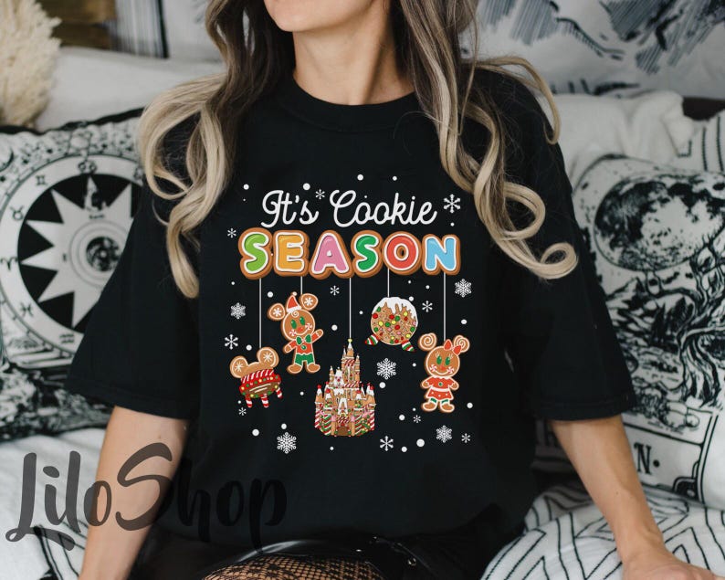 May include: Black t-shirt with the text "It's Cookie Season" in white and colorful letters. The shirt features gingerbread cookie designs, including a gingerbread house, gingerbread people, and snowflakes.