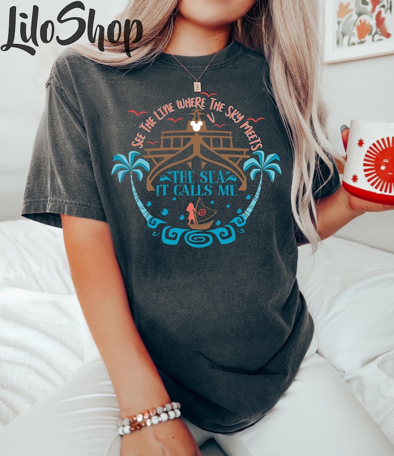 May include: Dark gray t-shirt featuring a circular design with the text "SEE THE LINE WHERE THE SKY MEETS THE SEA IT CALLS ME". The design includes a boat, palm trees, and a character illustration. The shirt is a comfortable fit.