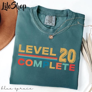 May include: A blue spruce Comfort Colors t-shirt with the text "LEVEL 20 COMPLETE" in yellow, red, orange, and blue. A gold necklace with a pendant is draped over the shirt. The shirt is folded.