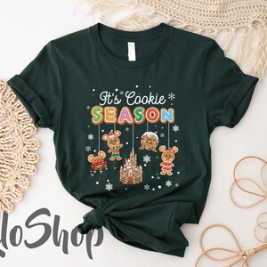May include: Dark green t-shirt with the text "It's Cookie Season" in colorful letters. The design includes gingerbread mouse characters, a gingerbread castle, and snowflakes, creating a festive holiday theme.