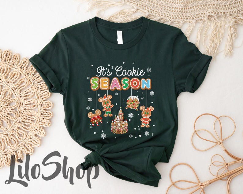 May include: Dark green t-shirt with the text "It's Cookie Season" in colorful letters. The design includes gingerbread mouse characters, a gingerbread castle, and snowflakes, creating a festive holiday theme.