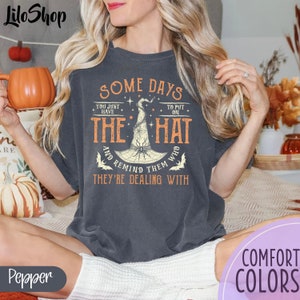 May include: A dark gray t-shirt with a graphic of a witch's hat and spider. The text on the shirt reads "Some days you just have to put on the hat and remind them who they're dealing with."