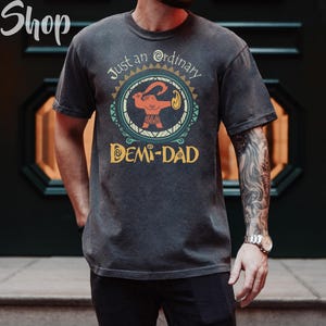 May include: Dark gray t-shirt with a graphic featuring a cartoon figure holding a weapon, surrounded by a circular design. Text on the shirt reads "Just an ordinary DEMI-DAD". The shirt has a vintage, worn look.