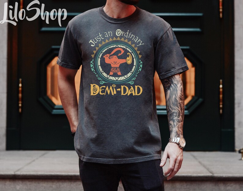 May include: Dark gray t-shirt with a graphic featuring a cartoon figure holding a weapon, surrounded by a circular design. Text on the shirt reads "Just an ordinary DEMI-DAD". The shirt has a vintage, worn look.