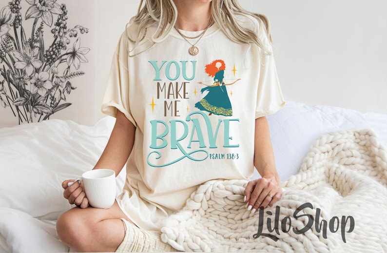 Może przedstawiać: Biały T-shirt z grafiką w kolorze turkusowym i złotym przedstawiającą kobietę z czerwonymi włosami. Grafika zawiera napis "You Make Me Brave" i "Psalm 138:3".
