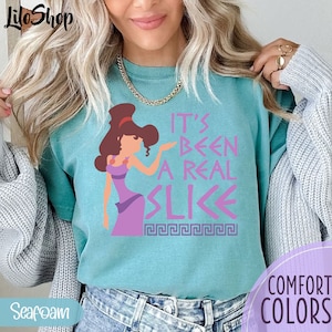 May include: A turquoise blue t-shirt with a pink graphic of a cartoon woman with brown hair and a purple dress. The text on the shirt reads "It's Been A Real Slice" with a Greek key design underneath.