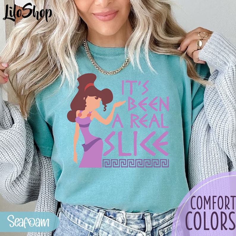 May include: A turquoise blue t-shirt with a pink graphic of a cartoon woman with brown hair and a purple dress. The text on the shirt reads "It's Been A Real Slice" with a Greek key design underneath.