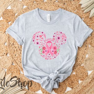 May include: A light gray short-sleeve t-shirt featuring a graphic in the shape of mouse ears. The design includes pink breast cancer awareness ribbons, flowers, and hearts, with the text Breast Cancer Hope printed in the center of the ribbon.