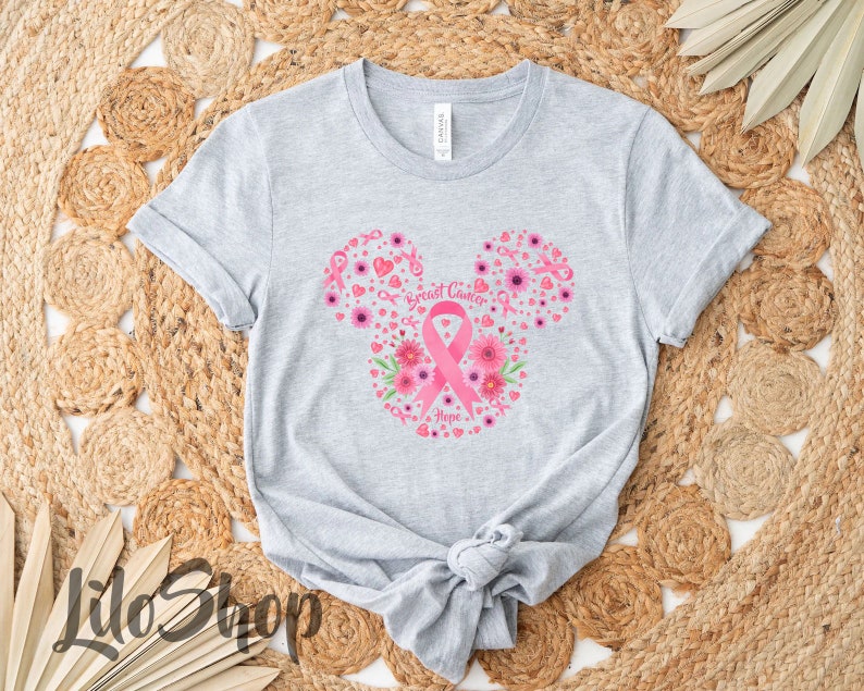 May include: A light gray short-sleeve t-shirt featuring a graphic in the shape of mouse ears. The design includes pink breast cancer awareness ribbons, flowers, and hearts, with the text Breast Cancer Hope printed in the center of the ribbon.