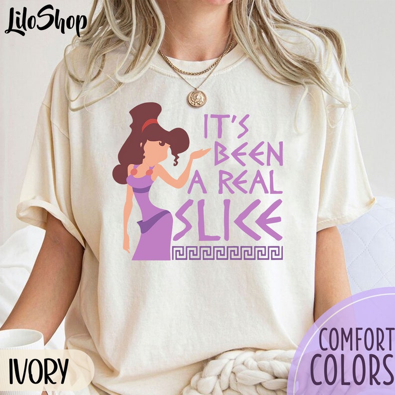 May include: A white t-shirt with a purple cartoon character and the text "It's been a real slice" in purple. The text is surrounded by a purple Greek key pattern.