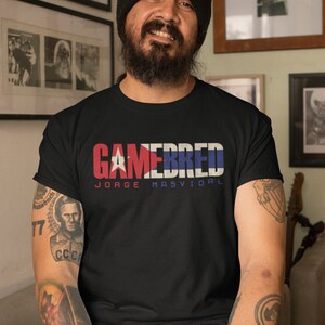 Puede incluir: Un hombre con un gorro negro y una camiseta negra con el texto "GAMEBRED JORGE MASVIDAL" en letras blancas. La camiseta tiene un diseño de bandera cubana. El hombre tiene barba y tatuajes en los brazos.
