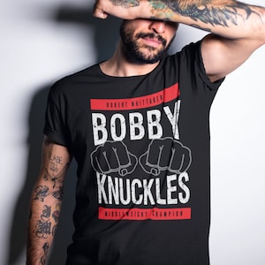 May include: Black t-shirt with a white graphic that says "Bobby Knuckles" with a red banner above and below that says "Robert Whittaker" and "Middleweight Champion".