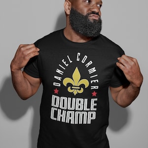 May include: Black t-shirt with a white graphic design featuring a fleur-de-lis, the text "DANIEL Cormier", two red stars, and the text "DOUBLE CHAMP".
