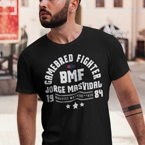 Puede incluir: Una camiseta negra con un gráfico circular blanco que dice "Gamebred Fighter BMF Jorge Masvidal 1984" con un pequeño gráfico de bandera estadounidense y tres estrellas.