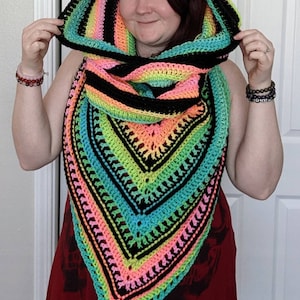 May include: A colorful crocheted hooded scarf with black, pink, green, and blue stripes. The scarf is worn by a person with red hair.