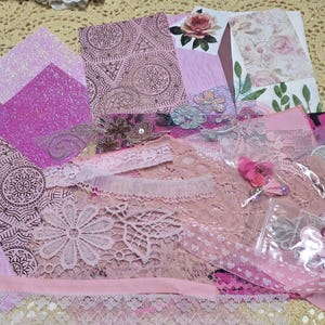 May include: A collection of pink and rose-colored craft supplies, including patterned paper, lace trim, and glitter paper. The assortment features various textures and floral accents, ideal for scrapbooking or crafting projects.
