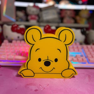 May include: A yellow Winnie the Pooh-shaped desk organizer with black outlines and a smiling face. The organizer is sitting on a pink surface, with a keyboard and other items in the background.