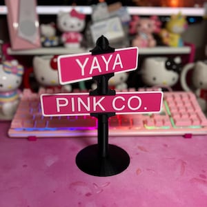 May include: A black plastic street sign stand featuring two rectangular pink signs with white borders and white text. The top sign reads YAYA and the bottom sign reads PINK CO. The stand has a circular base and a central pole holding both signs.