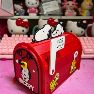 May include: A red mailbox-shaped decorative box with a Snoopy and Woodstock theme. The mailbox has a white flag that says "FOR YOU!" and features Snoopy and Woodstock designs. Other figurines are visible in the background.
