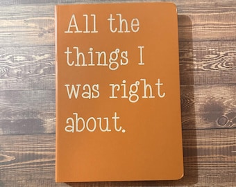 May include: An orange notebook with the text "All the things I was right about" in white. The notebook is rectangular and appears to be made of a smooth material. The background is a wooden surface.