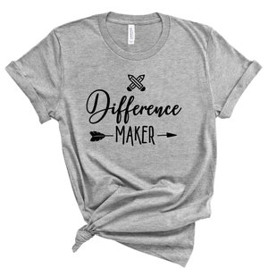 Op de afbeelding: Grijs T-shirt met een zwarte afbeelding met de tekst "Difference Maker" met een gekruist potloodontwerp en een pijl.